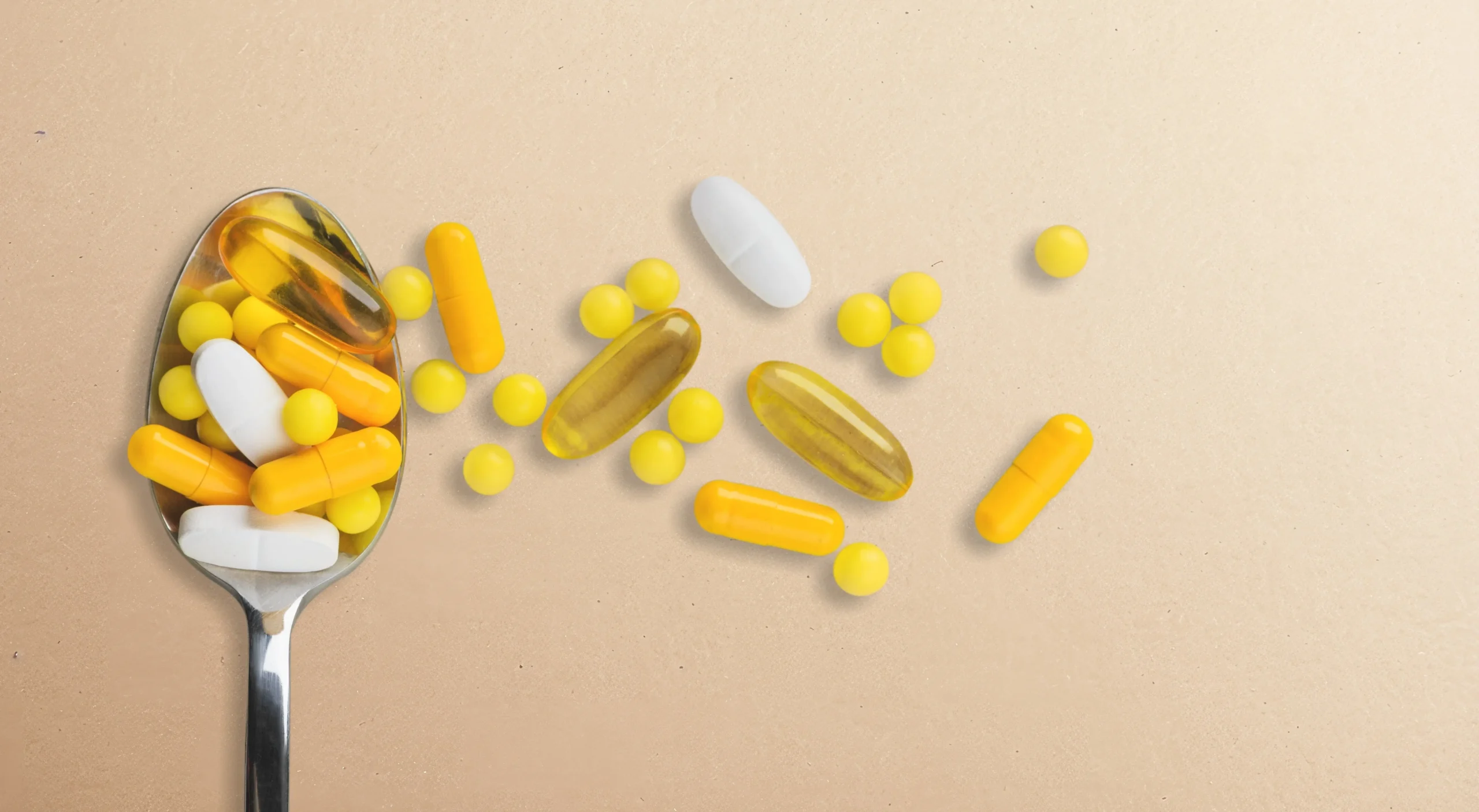 A metal spoon filled with various supplements including yellow capsules, white tablets, and small yellow round pills, with some pills scattered on a light beige surface.