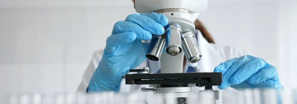 Laboratory scientist examining a sample through a microscope while wearing blue protective gloves in a third party product testing lab